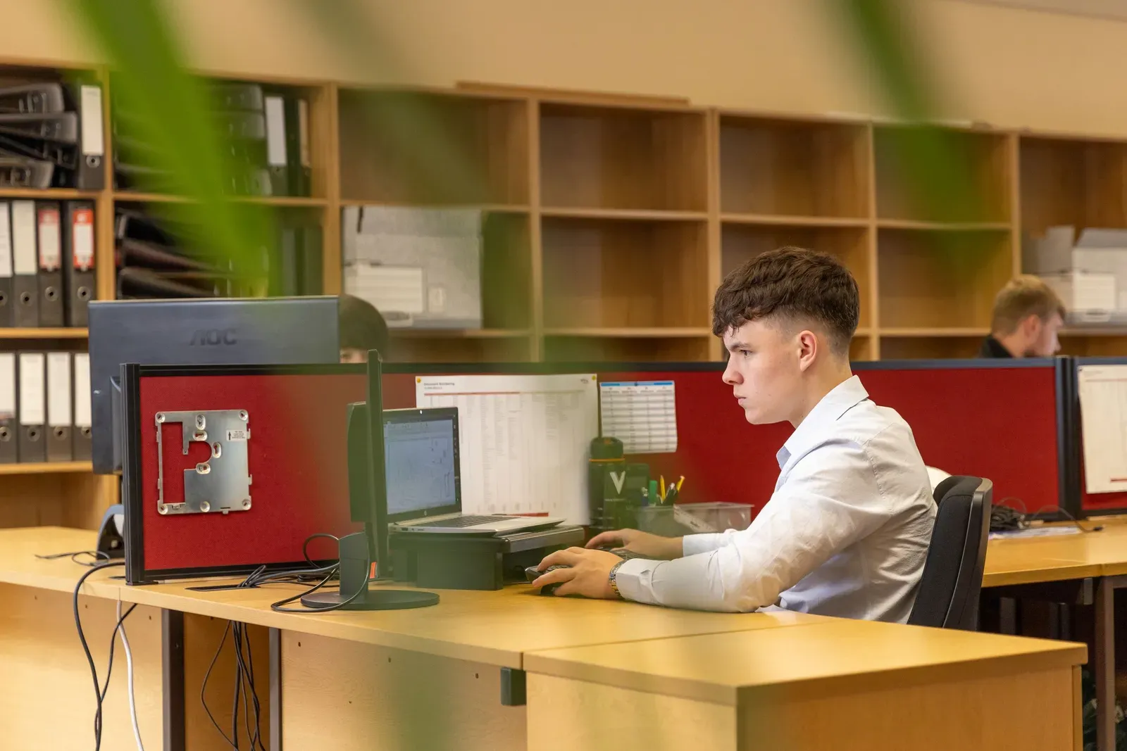 Medewerker achter een beeldscherm met een overzicht, met rijen ordners in de kast achter hem