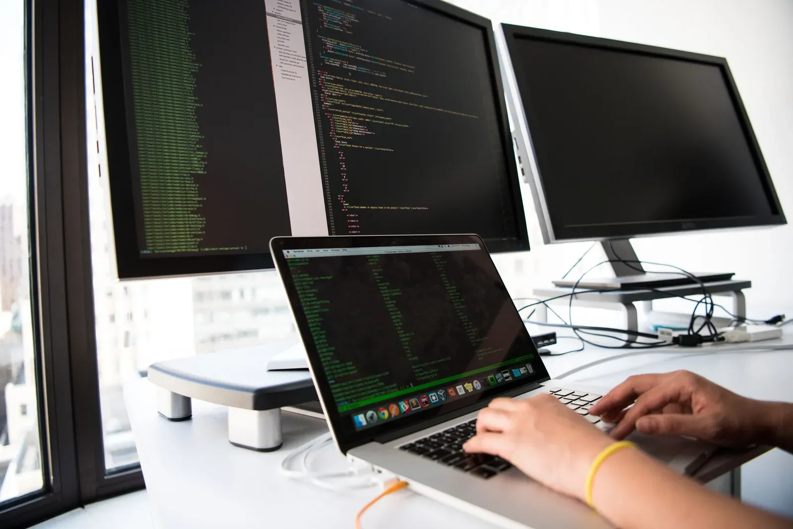 Laptop en twee beeldschermen met configuratiecode, handen typen op het toetsenbord