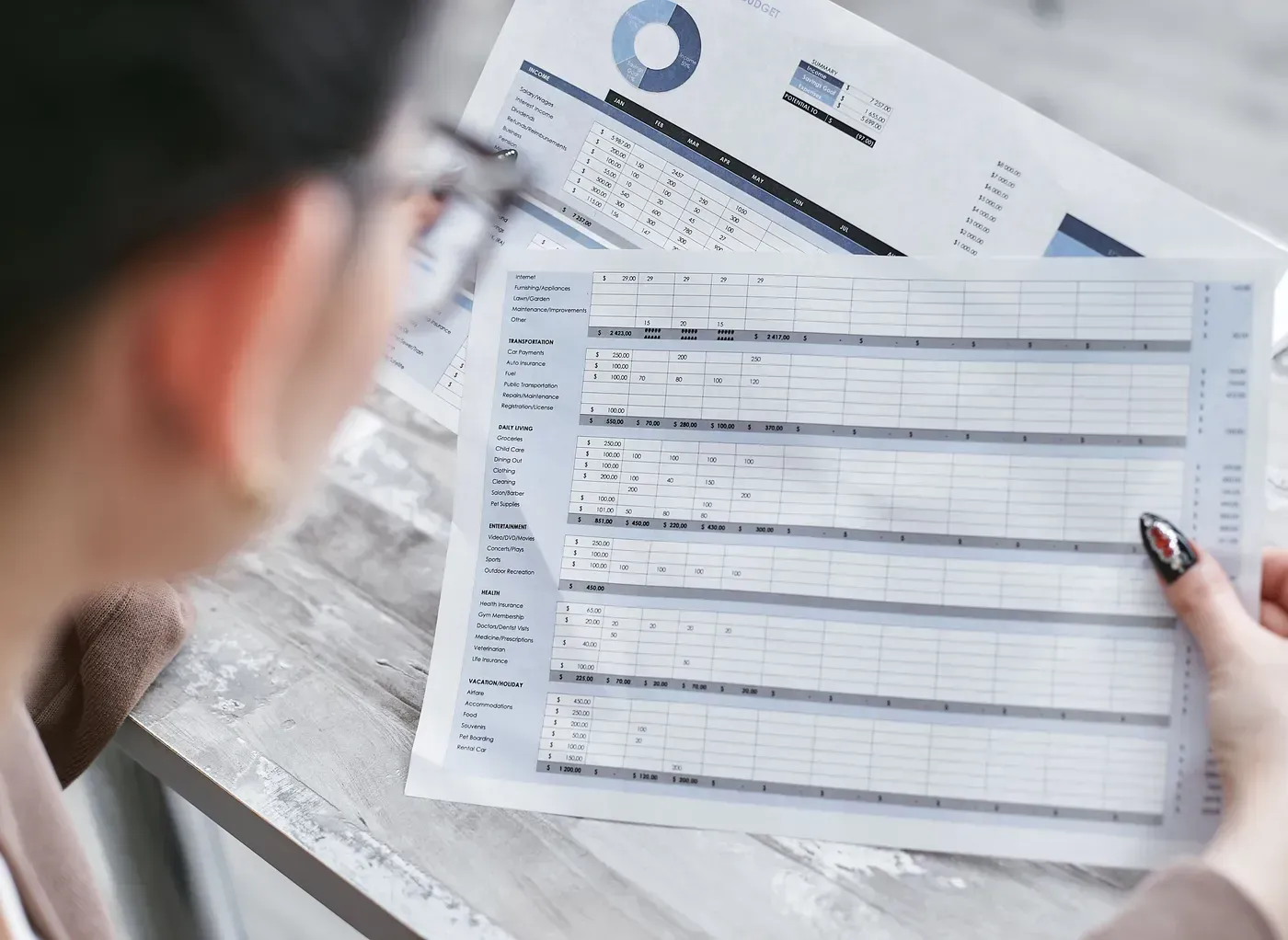 A woman holds two printed spreadsheets with figures side by side and compares them