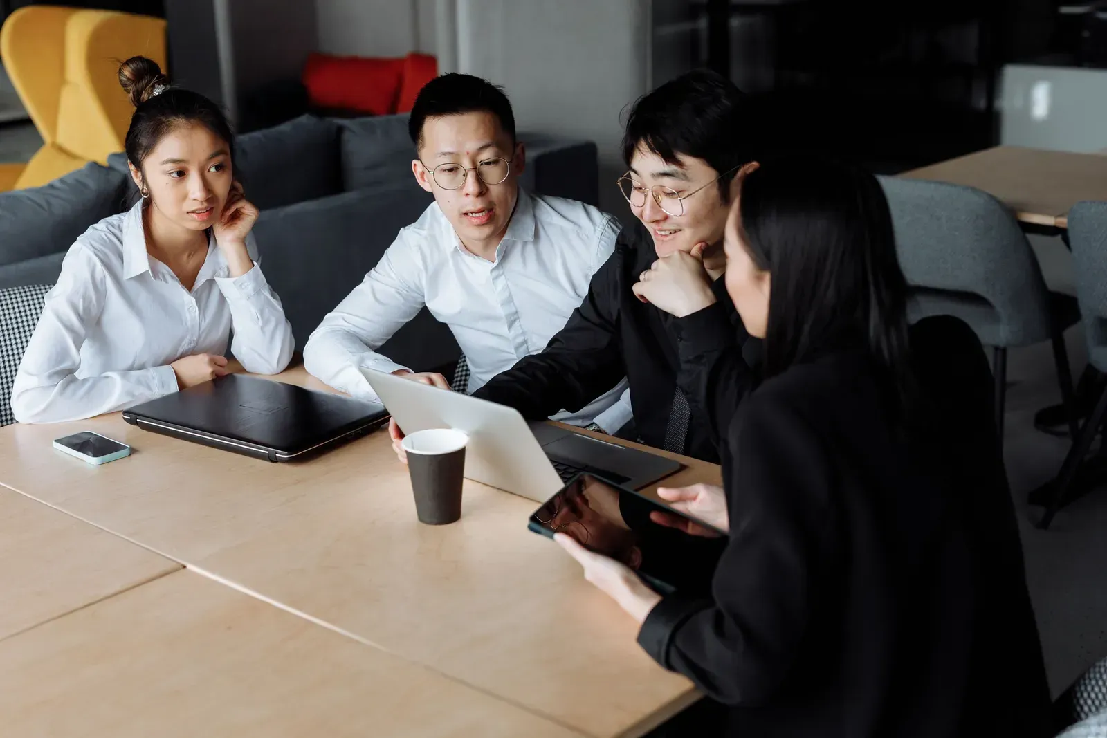 Four colleagues at a table look at a laptop and a tablet while one of them explains something