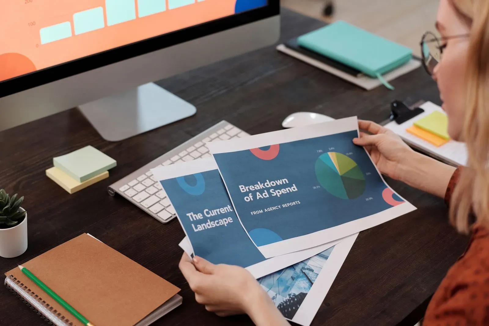 Vrouw aan een bureau bekijkt uitgeprinte rapporten, waarvan een met een cirkeldiagram en de titel Breakdown of Ad Spend