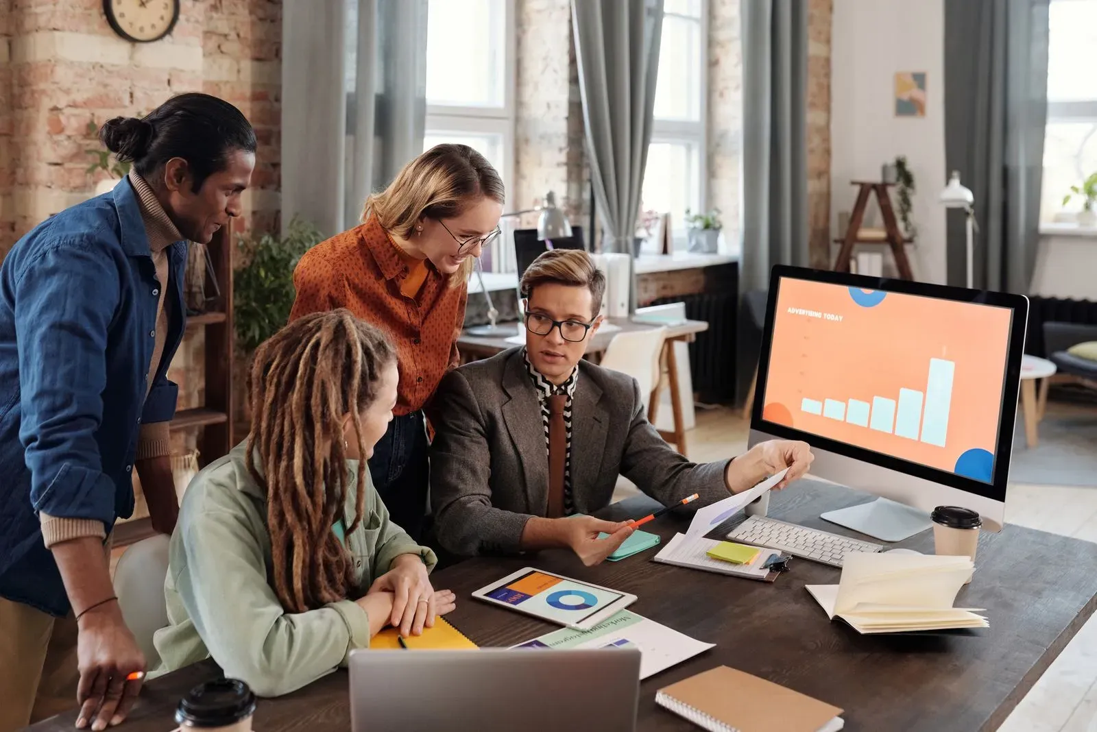 Vier collega's aan een bureau kijken mee naar een beeldscherm met een staafdiagram, met uitgeprinte grafieken en een tablet op tafel