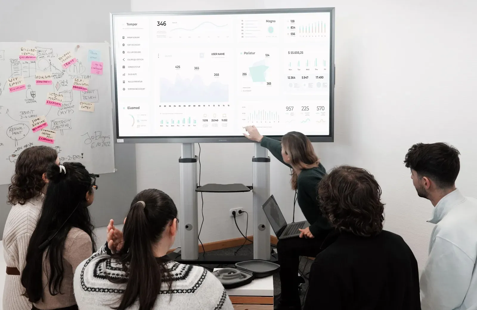 Team in a meeting room looking at a large screen showing a dashboard while a colleague points at a figure