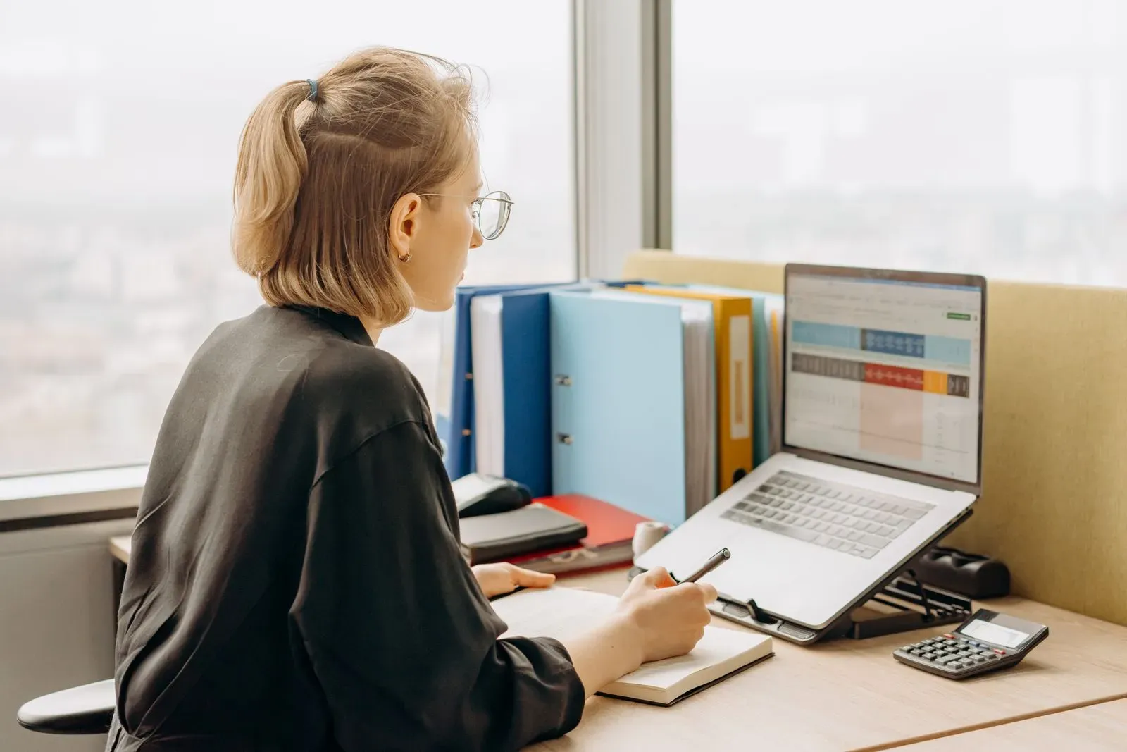 Woman wearing glasses looks at a spreadsheet on her laptop and takes notes in a notebook