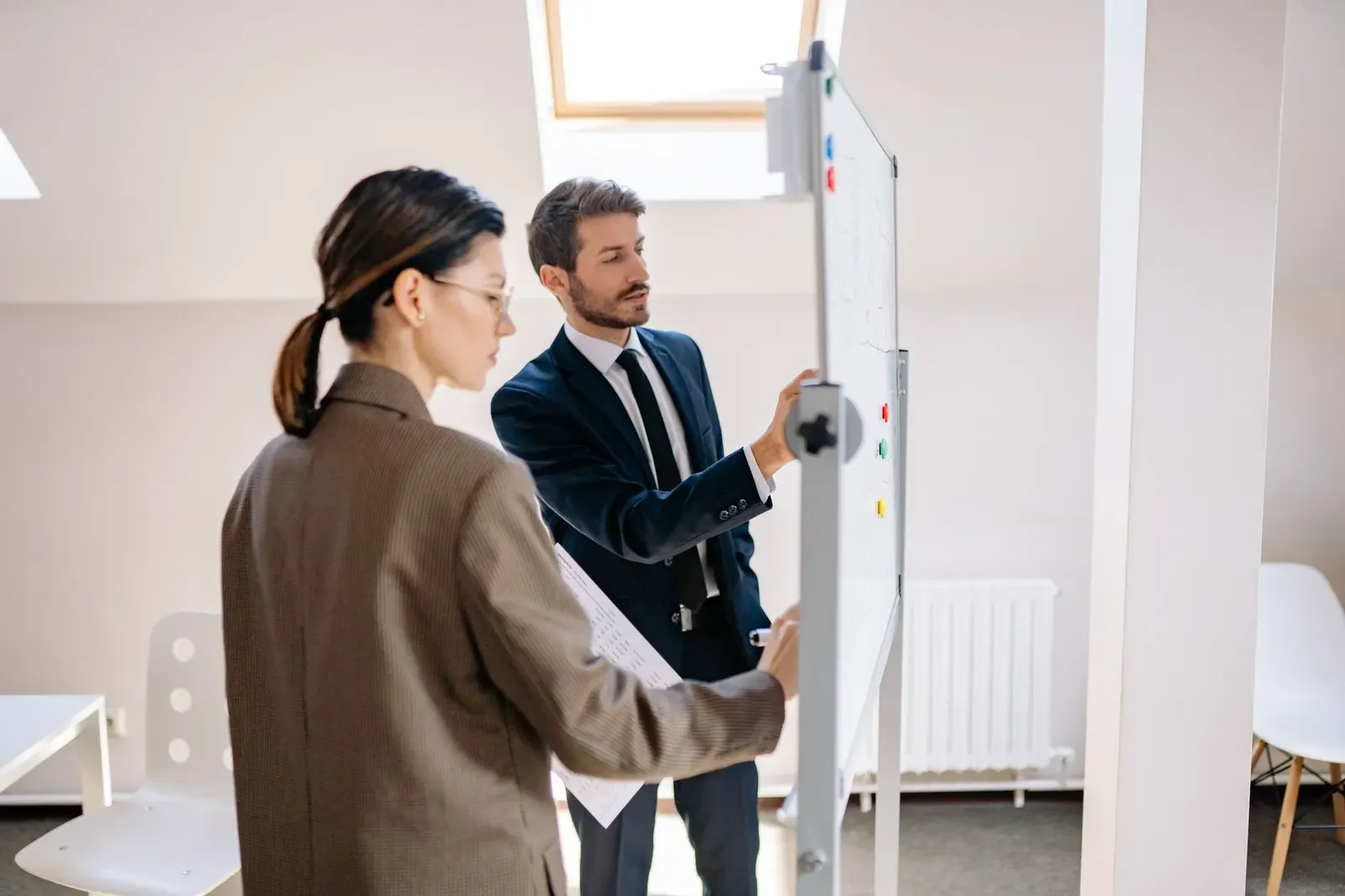 Een man in pak wijst iets aan op een whiteboard terwijl een collega met papieren in haar hand meekijkt