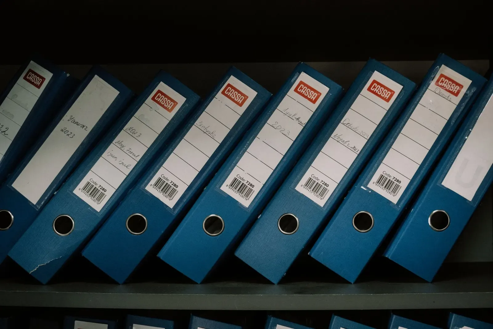 Rij blauwe ordners met handgeschreven etiketten en barcodes op een plank in een archiefkast