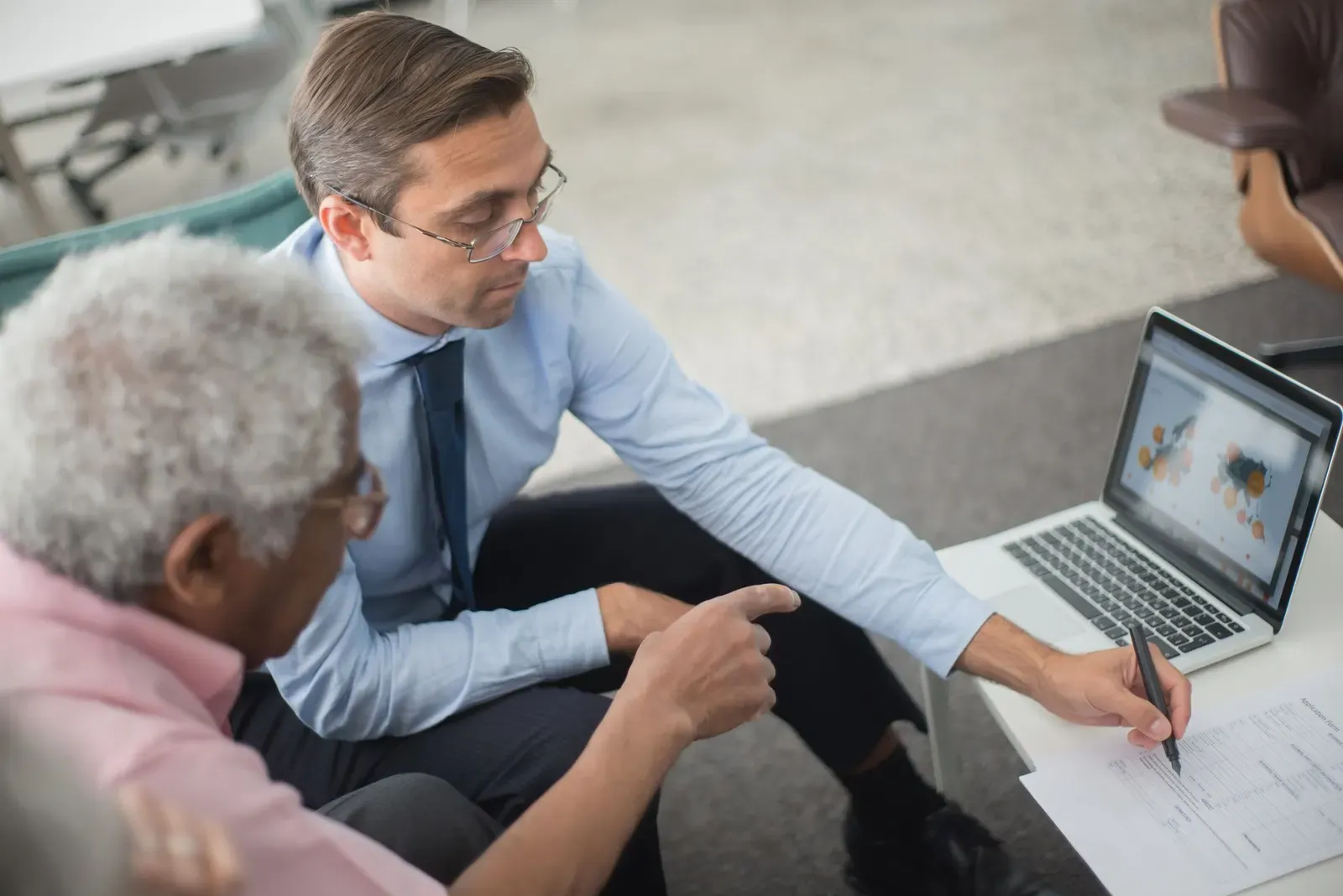 Twee mannen bespreken aan een tafel een document en een grafiek op een laptop, waarbij de een met zijn vinger iets aanwijst