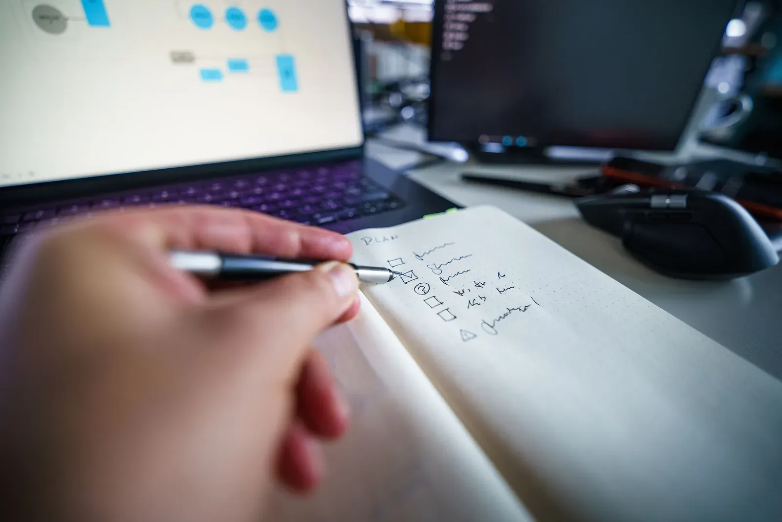 Hand schrijft een checklist in een notitieboek naast een laptop met een schema van gekoppelde systemen