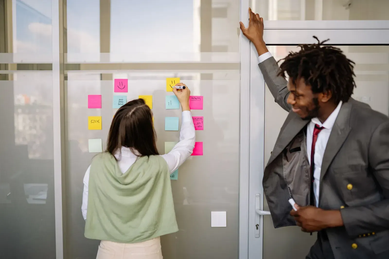 Vrouw schrijft op gekleurde memobriefjes die in een raster op een glazen wand hangen, terwijl een collega ernaast meekijkt