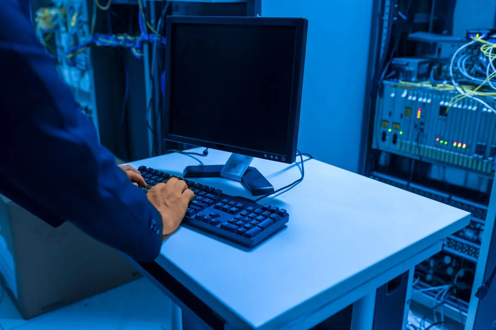 Someone typing at a desk keyboard in a server room with network equipment in blue light