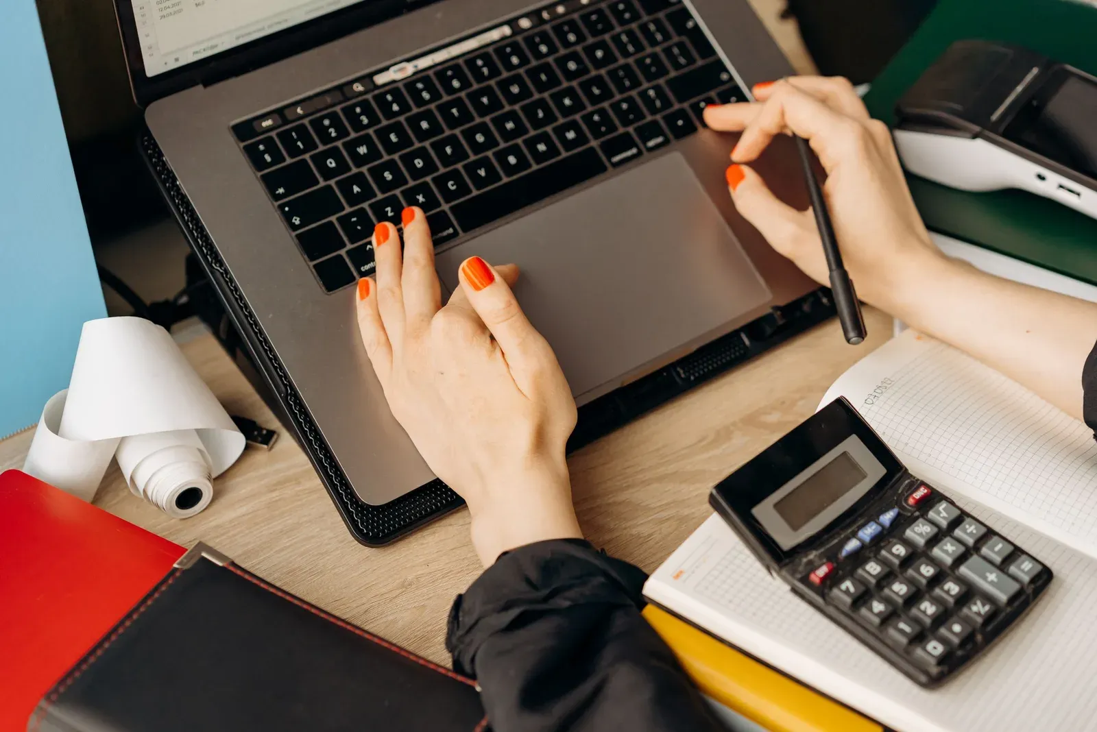 Hands on a laptop touchpad next to a calculator and a roll of receipt paper on a desk
