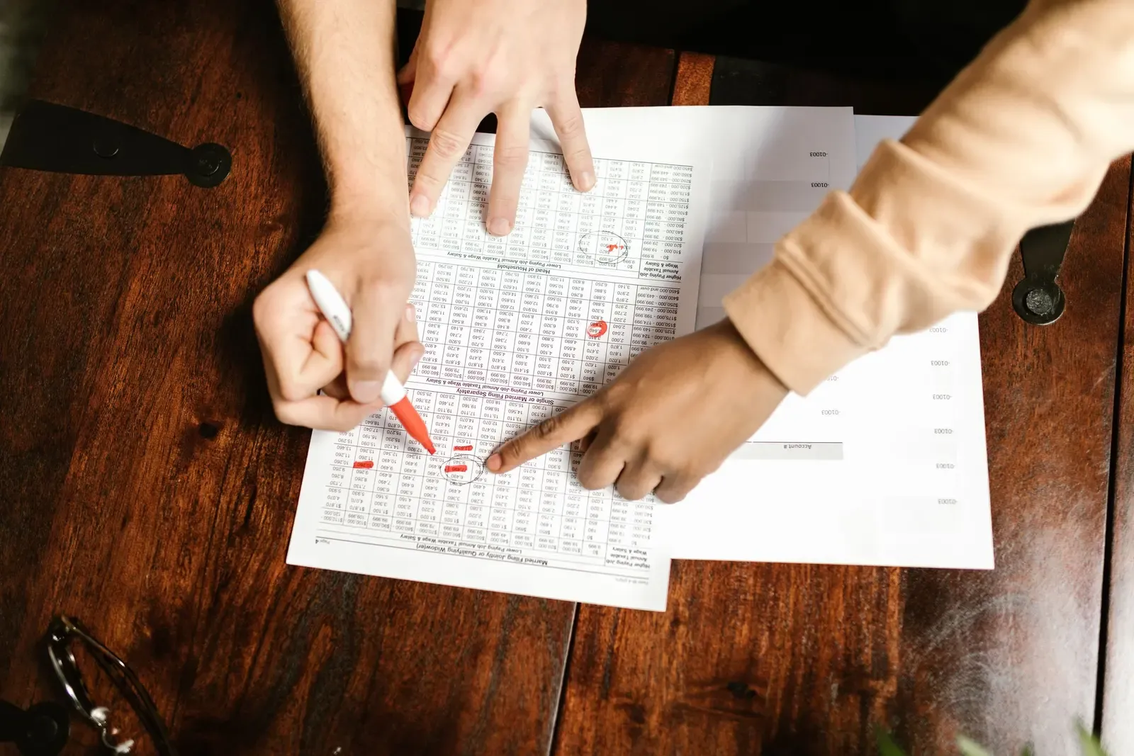 Two people pointing at lines in a printed table of amounts, one of them holding a red pen