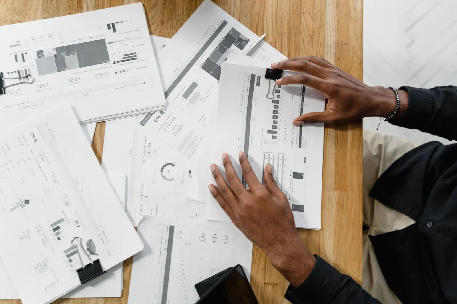 Desk covered in printed report pages full of charts and metrics, with someone sorting through them by hand