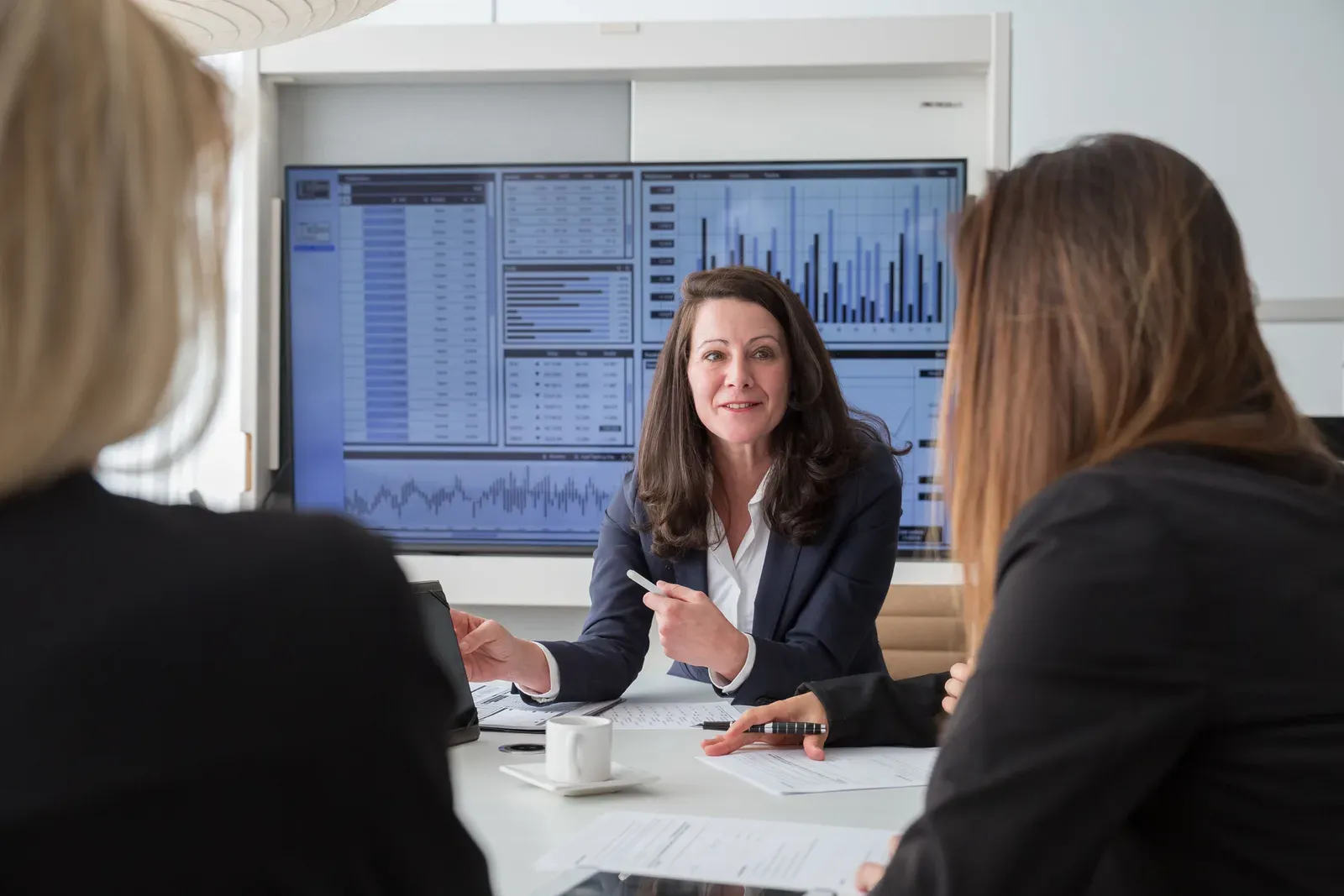 Management team in a meeting room discussing figures on a large screen showing tables and charts
