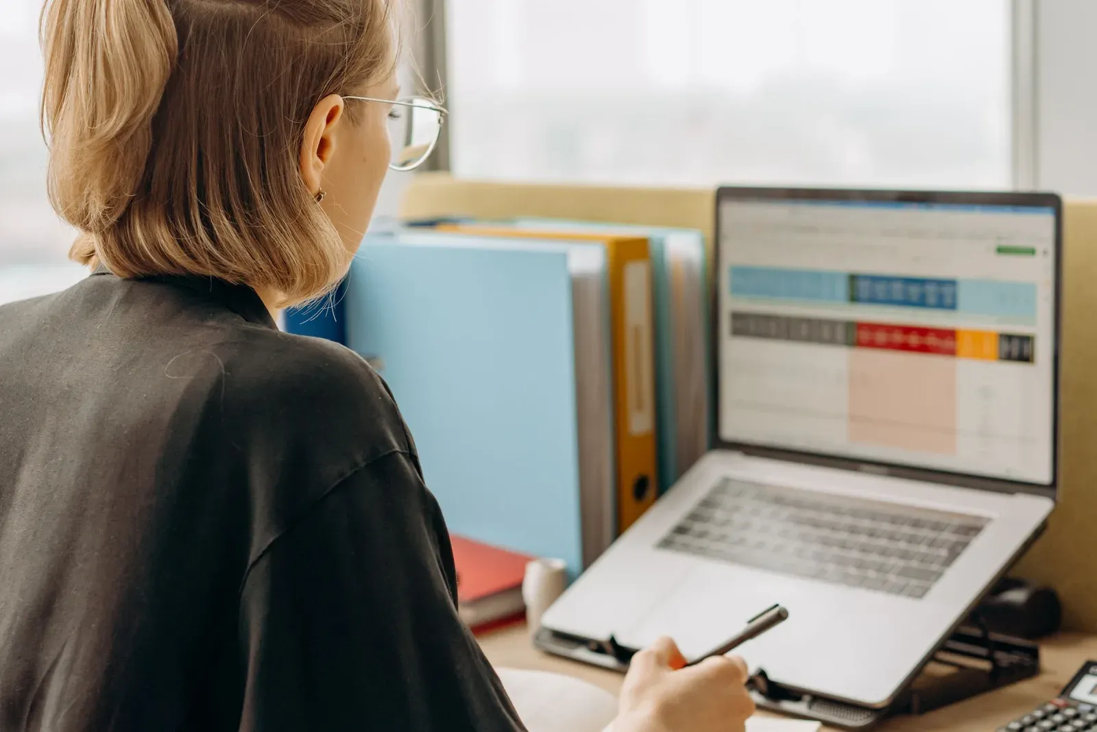 Vrouw met bril kijkt op een laptop naar een spreadsheet met blauwe, rode en oranje kolomkoppen, met ordners naast het scherm op haar bureau