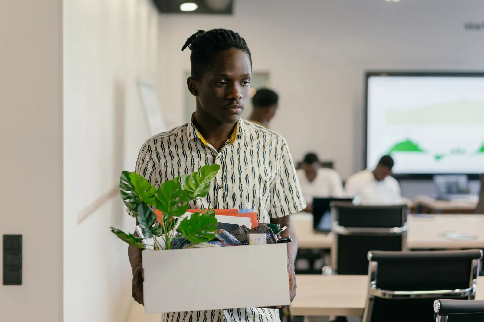 Man loopt met een doos vol spullen en een plant door een kantoor, met collega's aan hun bureau en een scherm met een grafiek op de achtergrond