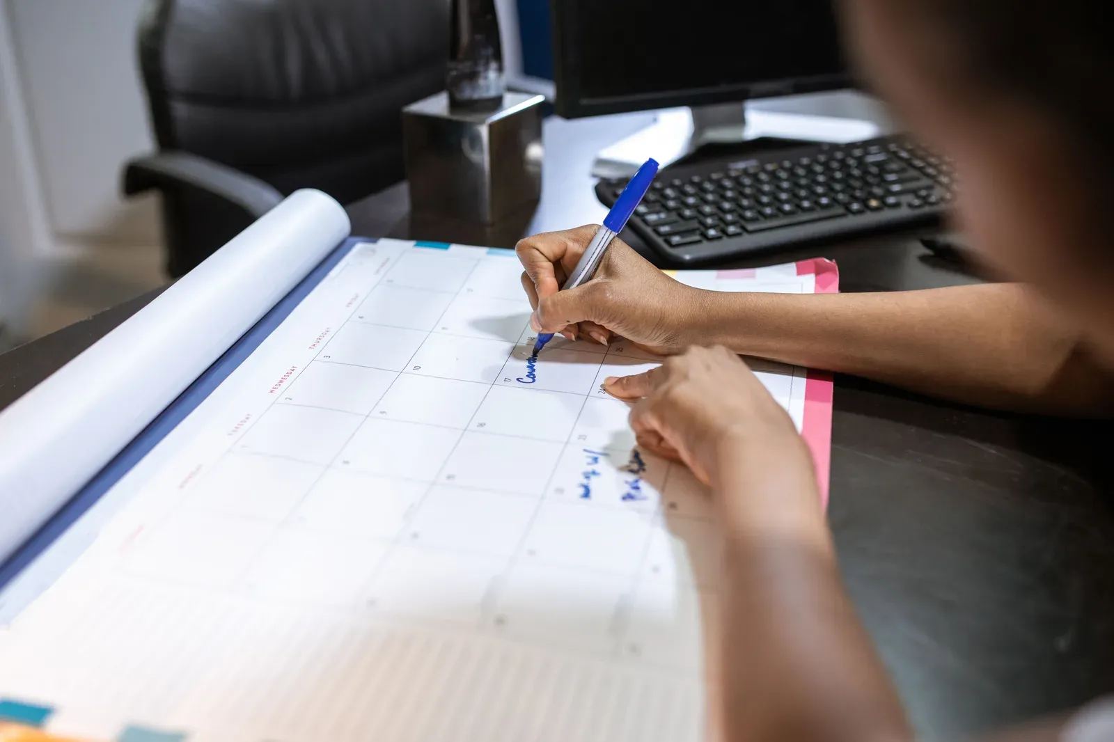 Handen schrijven met een pen een afspraak in een grote maandkalender op een bureau naast een toetsenbord