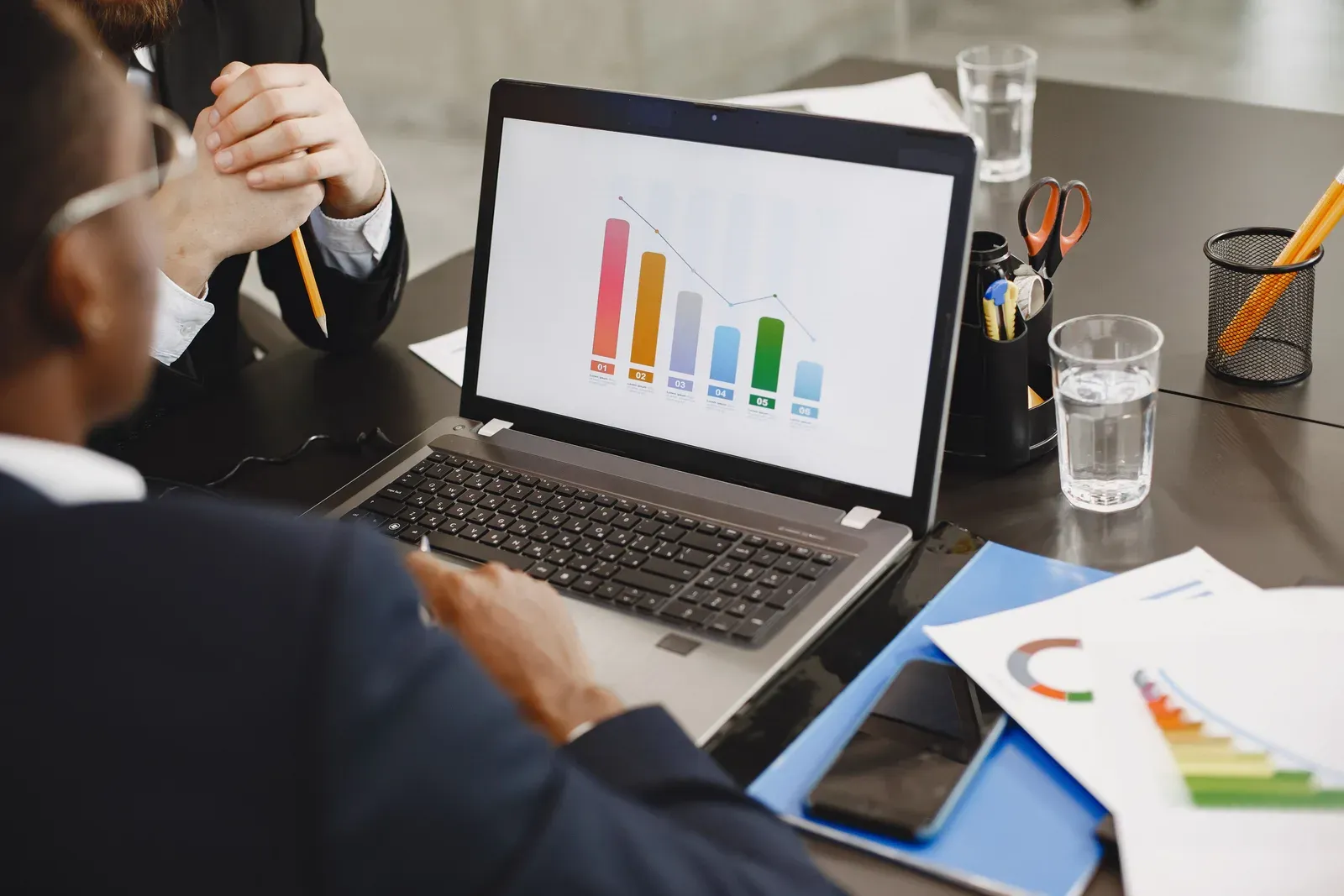 Twee collega's aan een vergadertafel kijken naar een laptopscherm met een staafdiagram, met uitgeprinte grafieken ernaast