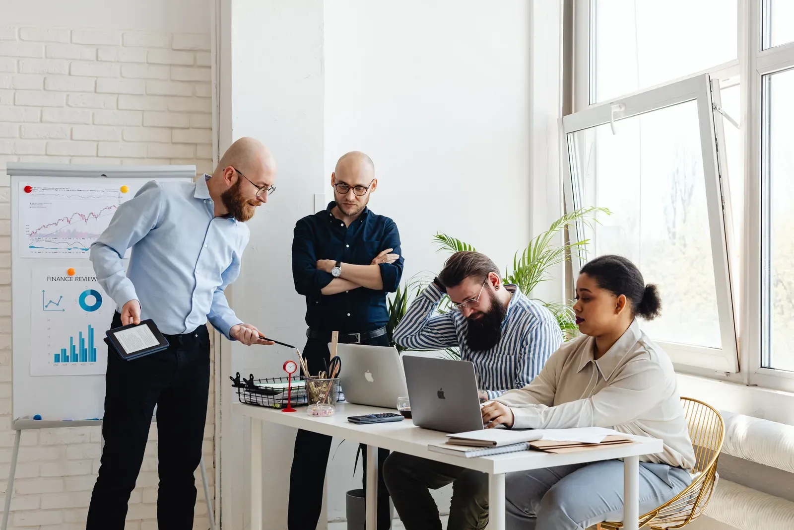 Vier collega's in een licht kantoor overleggen bij een bureau met laptops, naast een flip-over met staaf- en cirkeldiagrammen