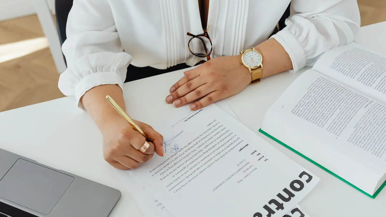 Vrouw ondertekent een contract aan een bureau, met een laptop en een opengeslagen boek ernaast