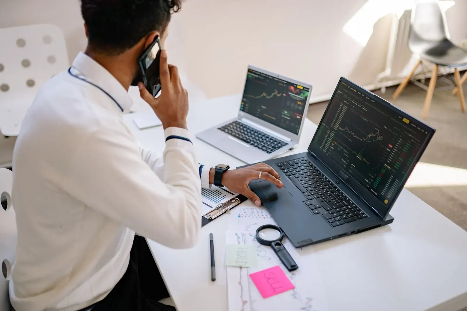 Man aan een bureau met twee laptops naast elkaar, op beide schermen grafieken, terwijl hij telefoneert