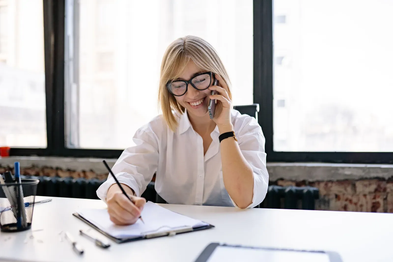 Vrouw aan een bureau voert een telefoongesprek en schrijft ondertussen aantekeningen op een klembord