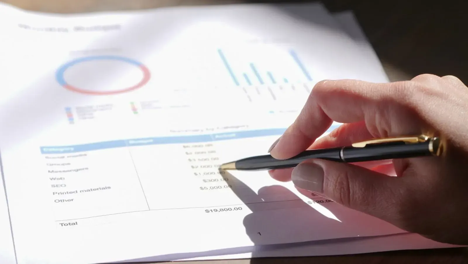 A hand holding a pen points at an item in a budget overview with columns for budget and actual